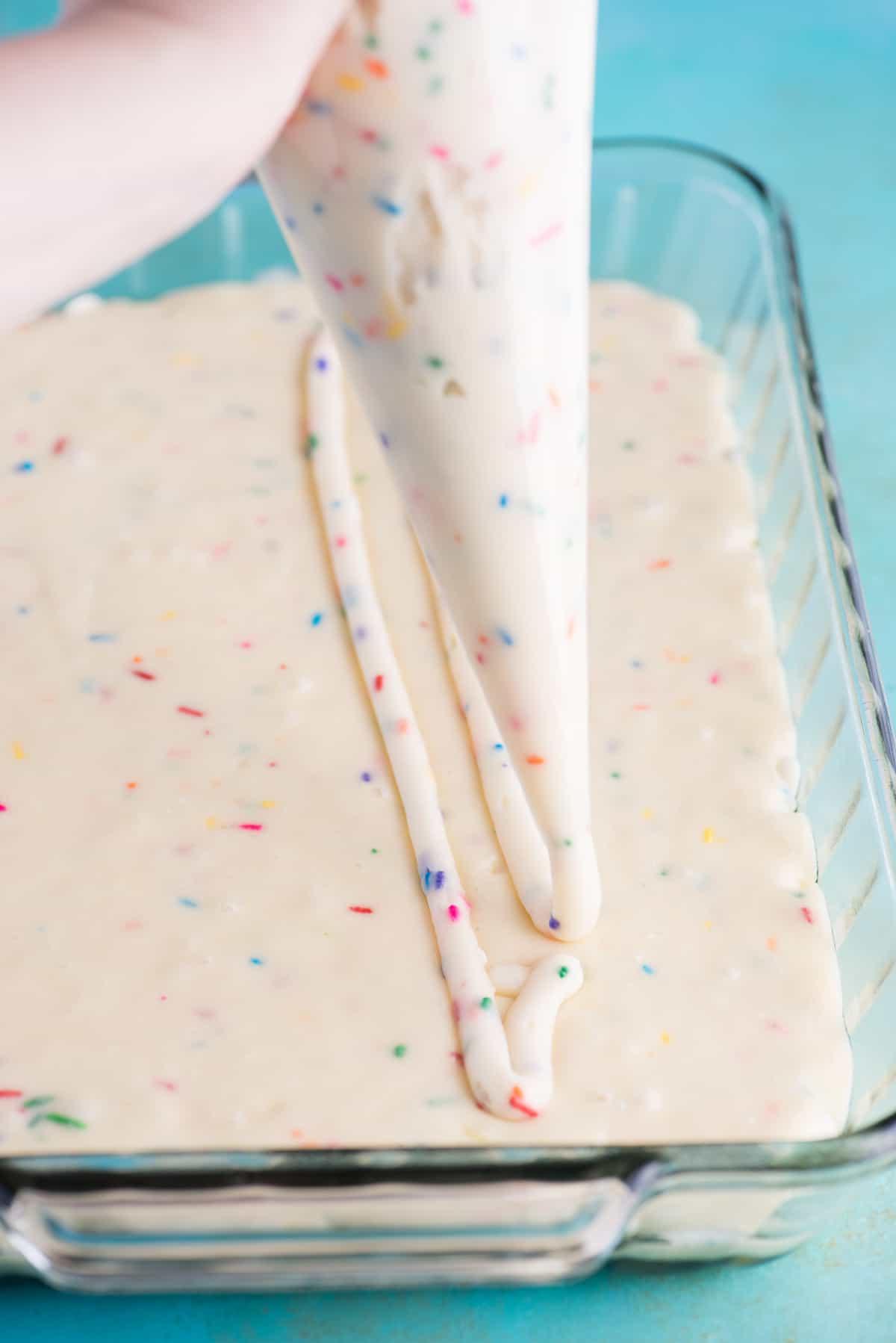 Questa torta terremoto funfetti è un mix per torta bianca arricchito con zuccherini e gocce di cioccolato funfetti. Lo strato di glassa al formaggio cremoso si fonde nella torta creando crepe e fessure in superficie. #earthquakecake #funfetticake #cake Glassa al formaggio cremoso disposta con sac à poche su impasto di torta bianca in teglia di vetro 9x13 pollici