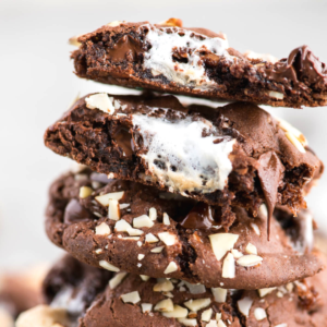 Primo piano di una pila di biscotti Rocky Road ripieni di marshmallow fusi e cioccolato, guarniti con noci tritate. Il biscotto superiore è spezzato a metà, rivelando il ripieno morbido all'interno.