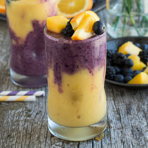 Frullato Hawaiano di Bacche 7 Due bicchieri di Smoothie Hawaiano ai Frutti di Bosco, con spirali gialle e viola, sono guarniti con fette di pesca, more e mirtilli su una superficie di legno rustica. Una cannuccia a strisce e un piatto di frutta completano la scena vibrante.