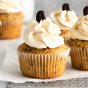 Cupcakes al caffè 7 Primo piano di Muffin al Caffè con glassa cremosa a vortice e un chicco di caffè al cioccolato sopra, posizionati su carta forno e una griglia di raffreddamento.