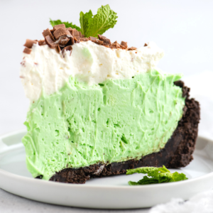 Torta Grasshopper Analcolica 7 Una fetta di torta grasshopper con una crosta di cioccolato, guarnita con panna montata, scaglie di cioccolato e un rametto di menta fresca, servita su un piatto bianco.