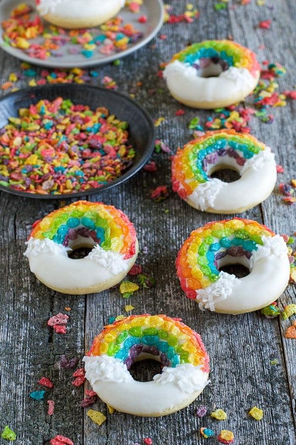 Ciambelle Arcobaleno - queste divertenti ciambelle sono fatte con cereali colorati, cioccolato bianco e cocco grattugiato! Non potrai fare a meno di queste deliziose ciambelle! Cinque Ciambelle Arcobaleno fatte in casa coperte di cioccolato bianco, scaglie di cocco e cereali Fruity Pebbles su un tavolo di legno.