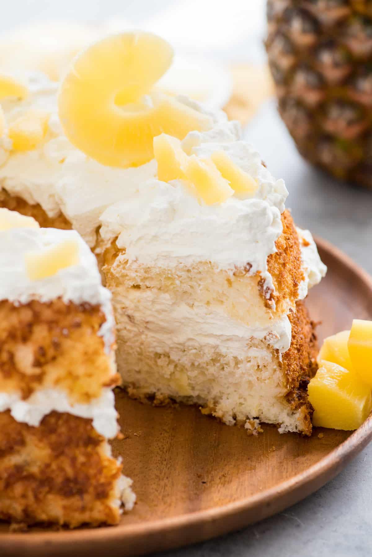 Torta Angel Food con fetta rimossa guarnita con panna montata e fette di ananas su vassoio di legno su sfondo bianco