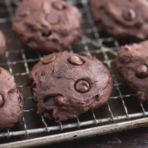 Biscotti Più Sani al Cioccolato Fondente e Olio di Cocco 3 Close-up of thick, soft healthier chocolate cookies with chocolate chips, cooling on a metal wire rack. The cookies appear rich, fudgy, and freshly baked.
