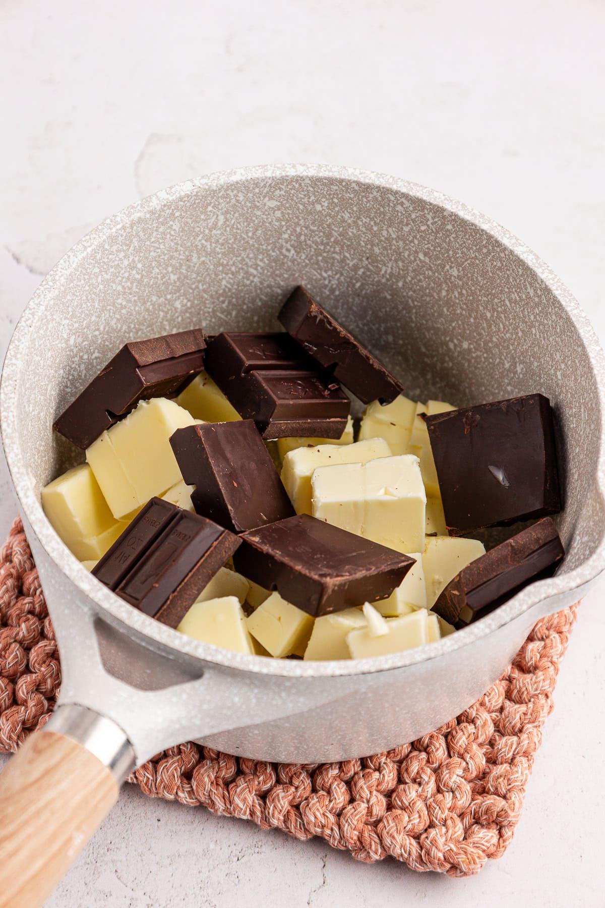 Una pentola contenente pezzi di cioccolato bianco e fondente è posta su un sottopentola rosa testurizzato sopra una superficie chiara.