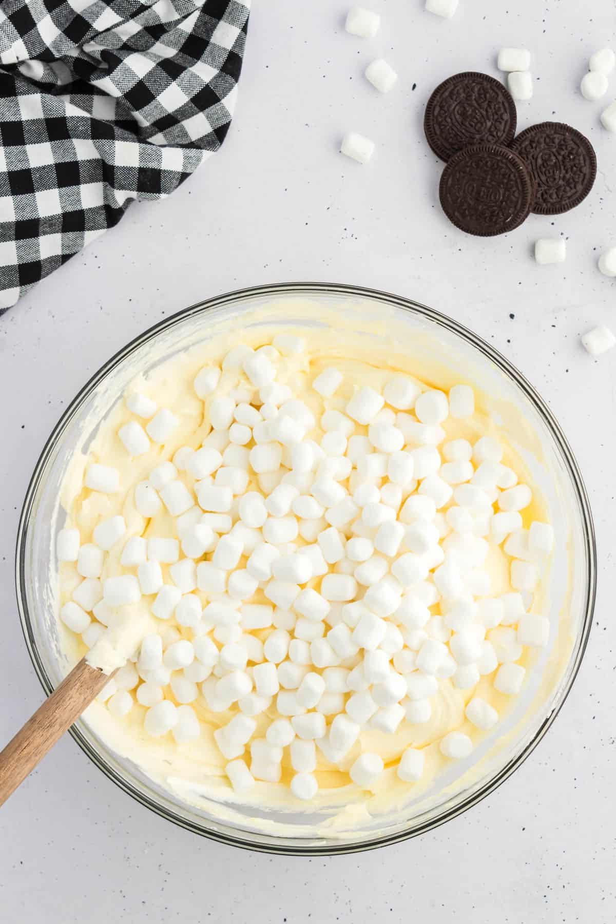 Ricetta Crema Soffice Oreo (Senza Formaggio Cremoso) 7 Miscela di budino alla vaniglia sormontata da mini marshmallow in una ciotola di vetro, circondata da Oreo, mini marshmallow e un panno da cucina a scacchi bianchi e neri