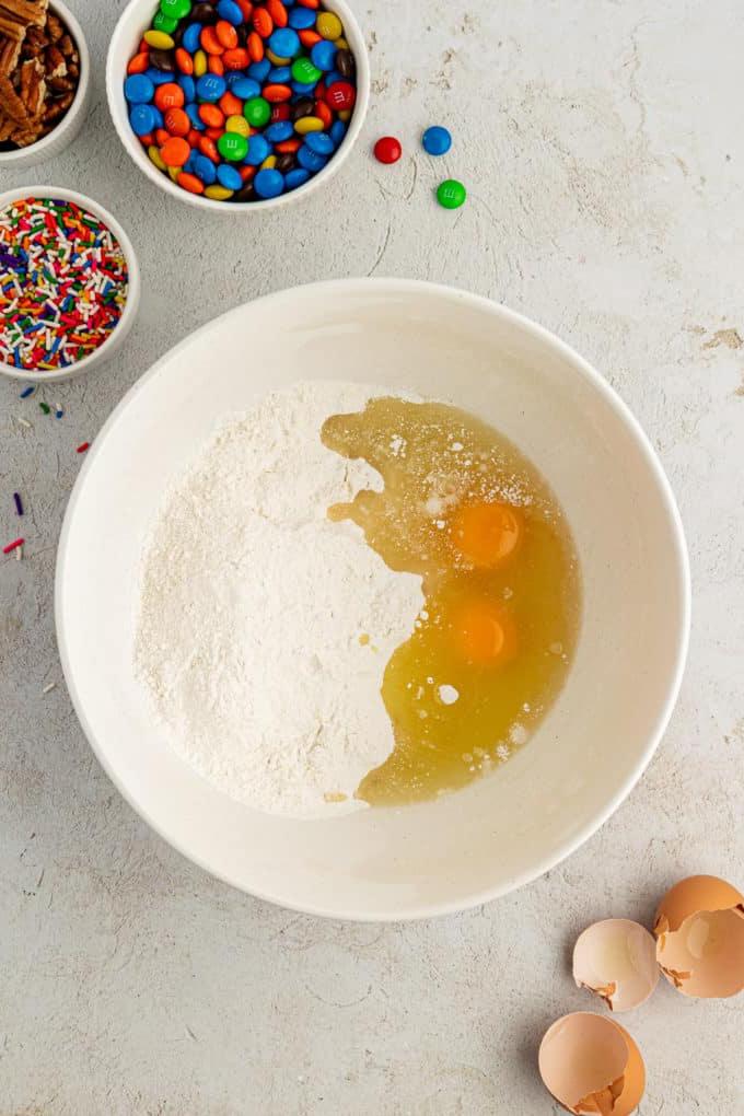 Fare Biscotti dal Mix per Torte 7 Uova e ingredienti secchi per biscotti con preparato per torte in una ciotola bianca circondata da gusci d'uovo, una ciotola di codette, una ciotola di M&M's e una ciotola di noci