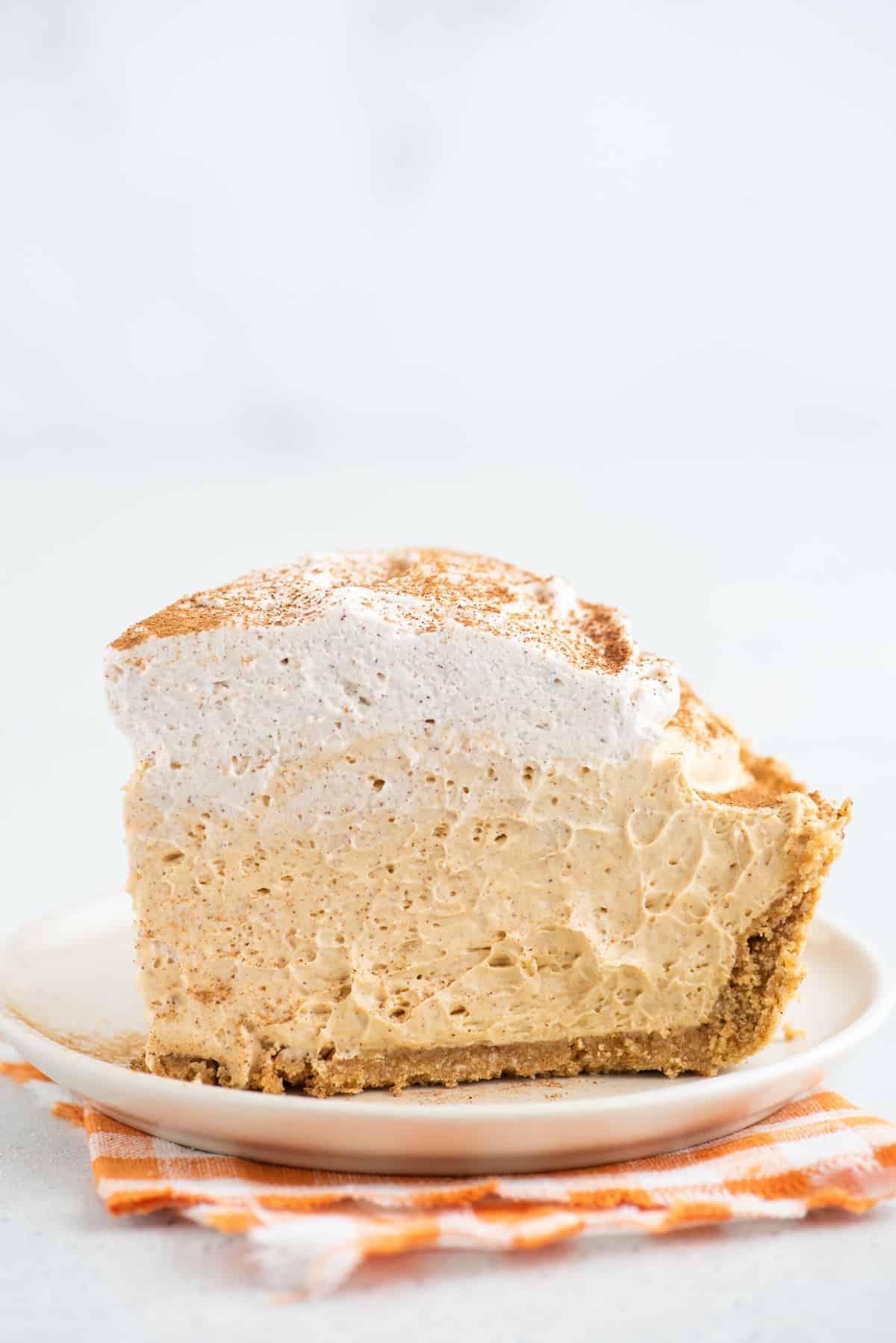 Questa torta di zucca senza cottura, veloce e facile, è leggera e cremosa - una deliziosa variante della classica torta di zucca. Una fetta alta di torta di zucca senza cottura è adagiata su un piatto bianco.