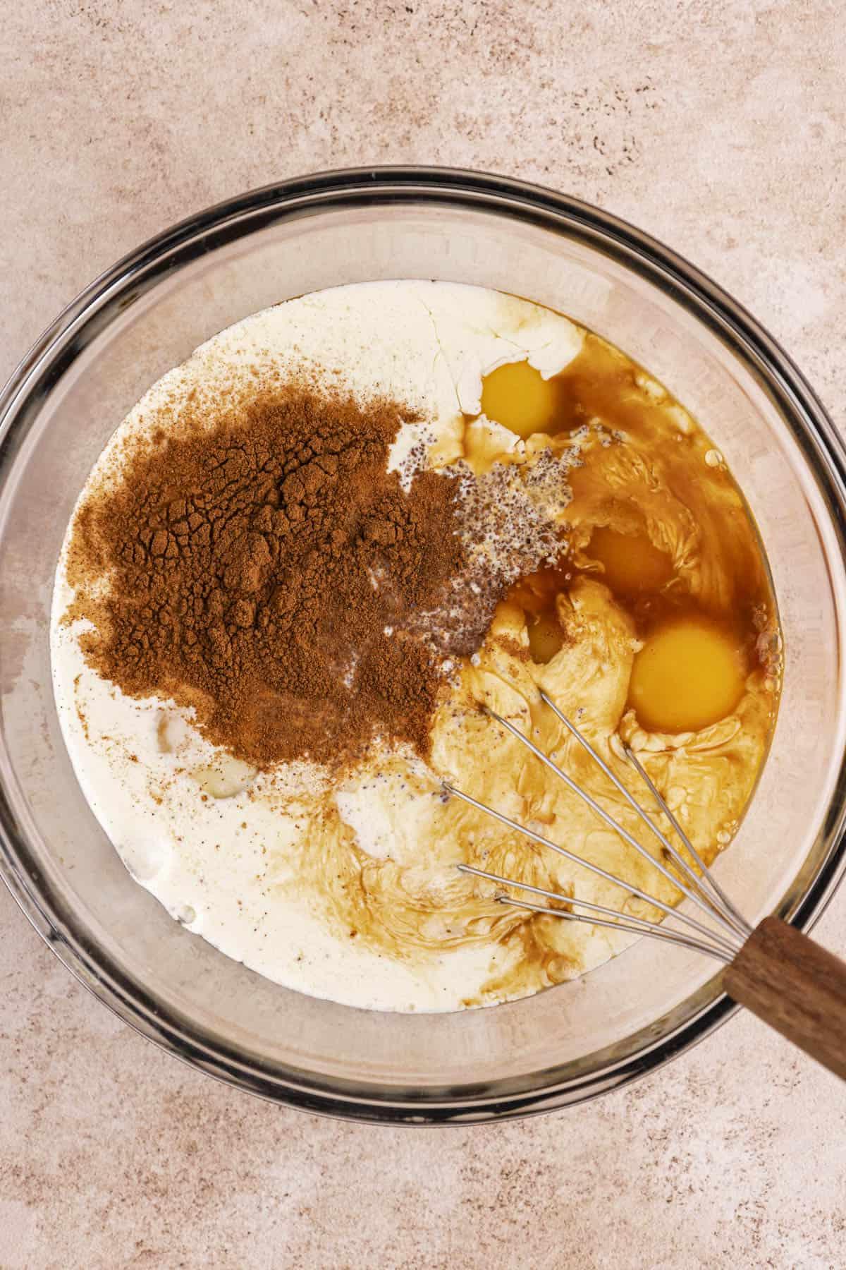 Migliore Ricetta per Budino di French Toast 7 Una ciotola di vetro trasparente con uova, latte intero, panna fresca, zucchero di canna, cannella, sale ed estratto di vaniglia con una frusta appoggiata all'interno della ciotola.
