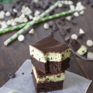 Brownies con Mousse alla Menta e Cioccolato 7 Tre brownies al cioccolato impilati con uno strato cremoso di menta al centro sono posti su un pezzo di carta forno. Gocce di cioccolato bianco e fondente e cannucce verdi sono sparse su una superficie di legno sullo sfondo.