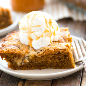 Una fetta di torta terremoto alla zucca umida e dorata con una pallina di gelato alla vaniglia irrorata di salsa al caramello, servita su un piatto bianco con una forchetta.
