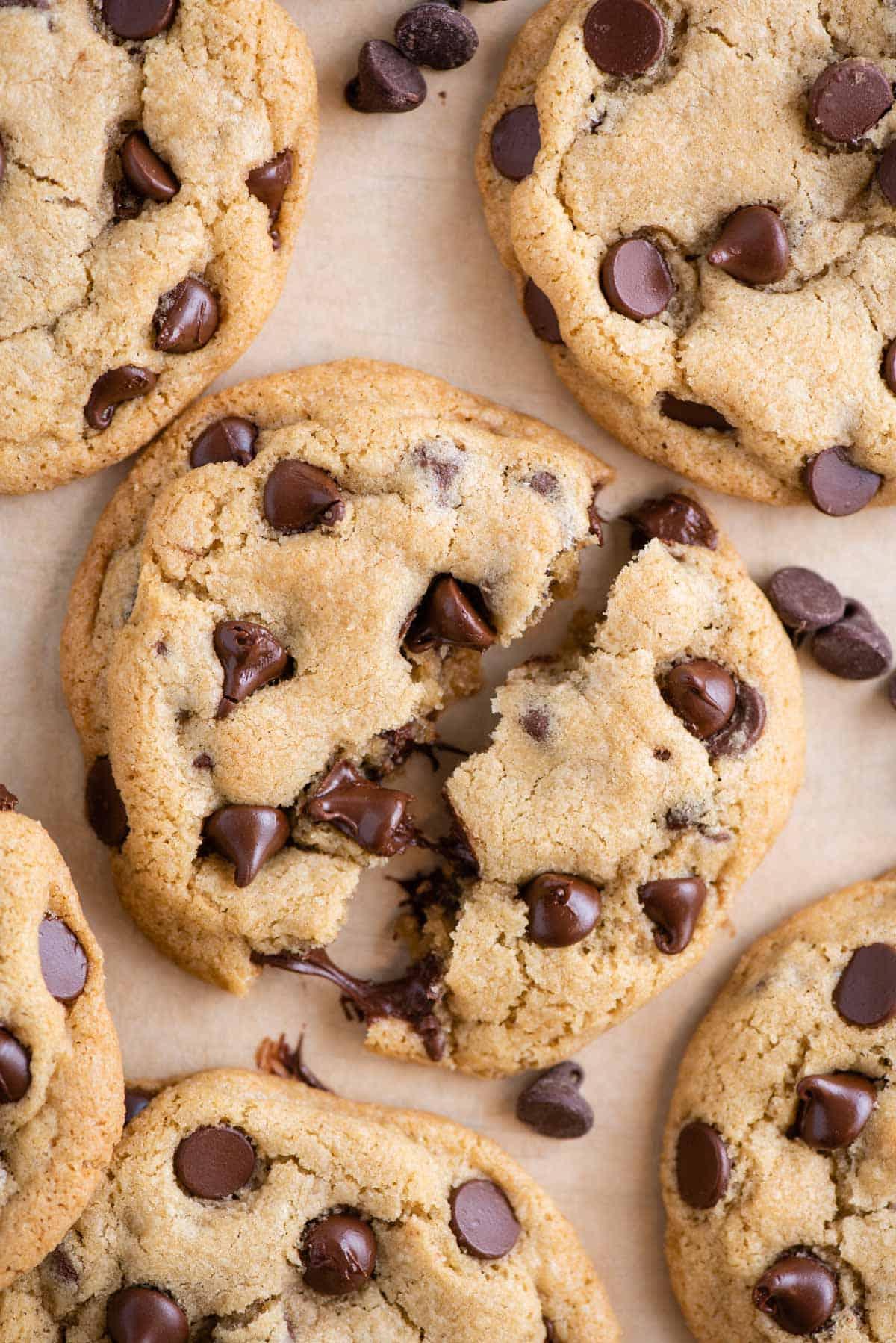 Se ami i biscotti morbidi e gommosi, adorerai questi biscotti con gocce di cioccolato in piccola quantità. Ne produce 6, perfetti per soddisfare una voglia! Visuale dall'alto di biscotti con gocce di cioccolato su carta forno, con uno spezzato a metà per mostrare le gocce di cioccolato fondenti