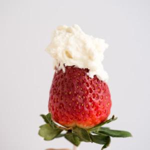 Cheesecake Panna Montata 2 Una mano che tiene una fragola fresca su uno stecchino, guarnita con una generosa cucchiaiata di panna montata al cheesecake su uno sfondo chiaro e semplice.