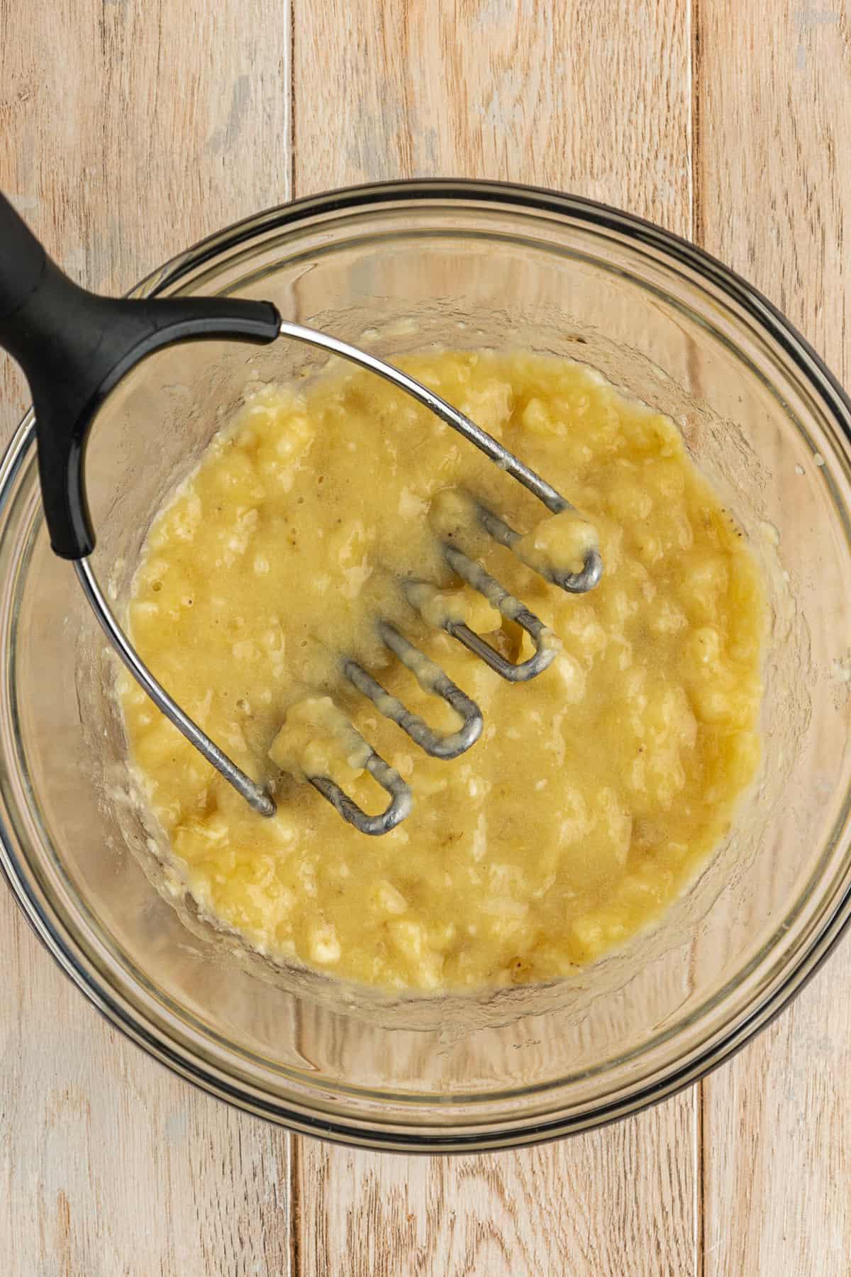 Ricetta Facile Torta alla Banana 7 Una ciotola di vetro piena di banane schiacciate con uno schiacciapatate appoggiato nella ciotola, su una superficie di legno