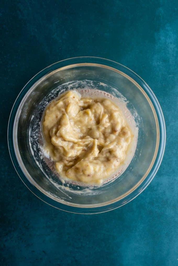 Il Miglior Pan di Banane con Gocce di Cioccolato 7 Banane schiacciate in una ciotola di vetro trasparente su una superficie color ottanio scuro.
