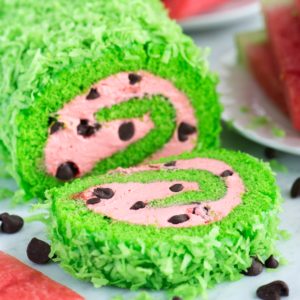 Una deliziosa torta rotolo anguria decorata per sembrare un'anguria, con scaglie di cocco verde brillante all'esterno, ripieno rosa a spirale, e gocce di cioccolato che ricordano i semi.