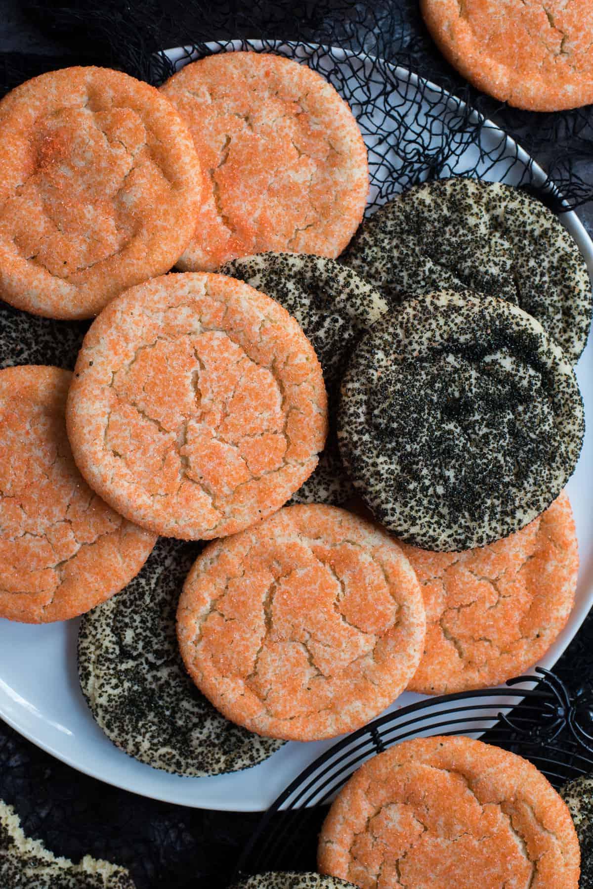 Semplici Biscotti di Zucchero di Halloween 7 Biscotti di zucchero arancioni e neri ammucchiati su piatto bianco con tessuto nero su sfondo nero