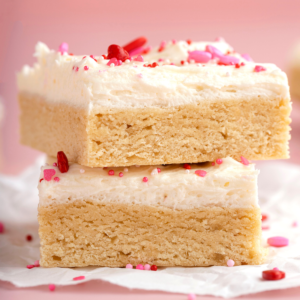 Quadrotti di Biscotti di Zucchero di San Valentino 8 Due spesse barrette di biscotto zuccherato di San Valentino impilate, guarnite con glassa bianca cremosa e decorate con confettini rosa, rossi e bianchi, poggiate su carta forno accartocciata su uno sfondo rosa—perfette per idee di biscotti di San Valentino.