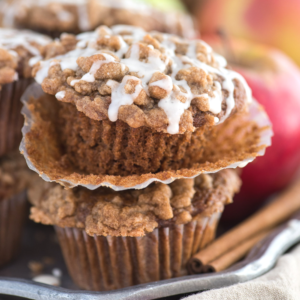 Due muffin alle mele con crumble, guarniti con streusel di cannella croccante e una pioggia di glassa, con un muffin parzialmente scartato. Una mela e bastoncini di cannella sono sullo sfondo.