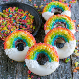 Ciambelle Arcobaleno 2 Cinque ciambelle arcobaleno decorate con cereali color arcobaleno e glassa bianca che assomiglia a nuvole sono disposte su una superficie di legno rustica, con una ciotola di pezzi di cereali colorati nelle vicinanze.