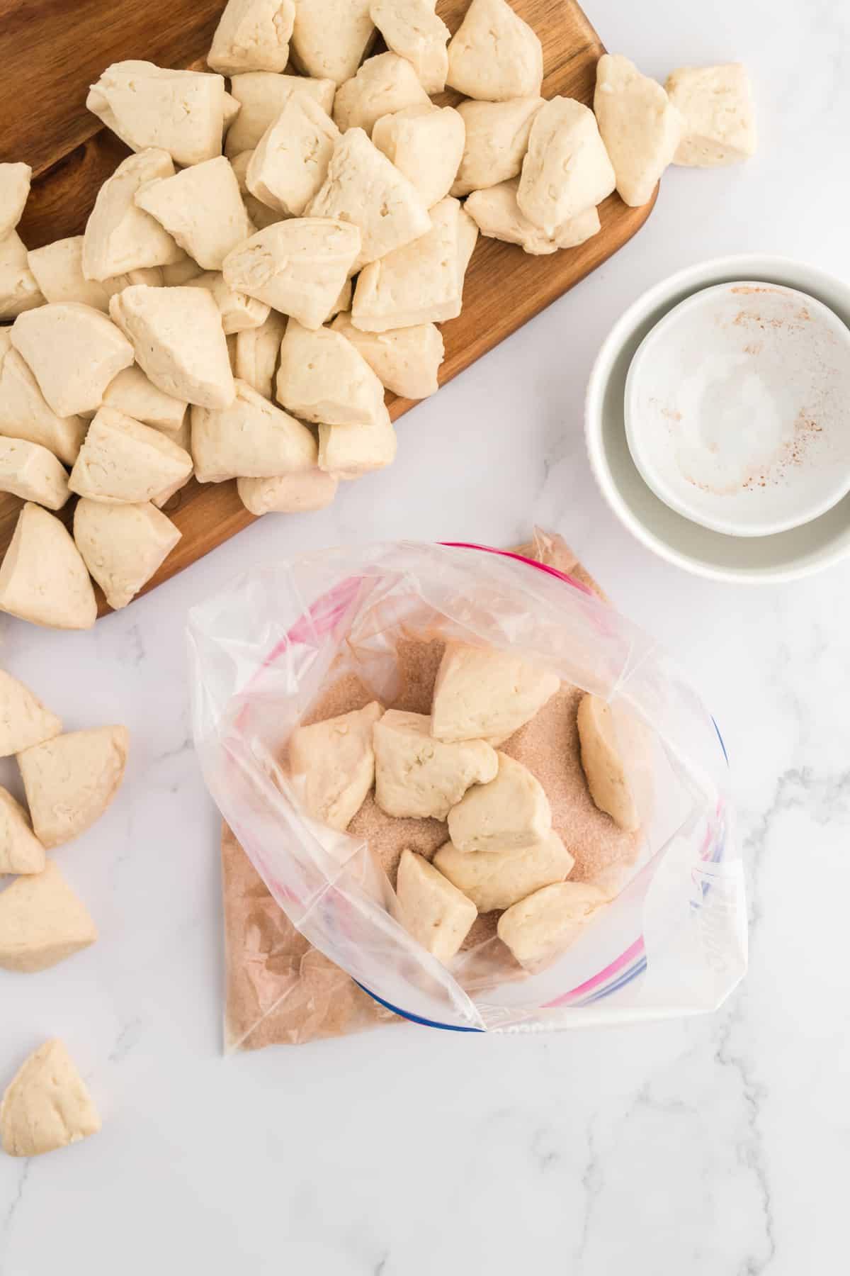 Pane delle scimmie di biscotti 8 Impasto di biscotti tagliato a pezzi ammassato su un tagliere accanto a una busta di plastica da un gallone di pezzi di impasto di biscotti e con una miscela di zucchero alla cannella sul fondo