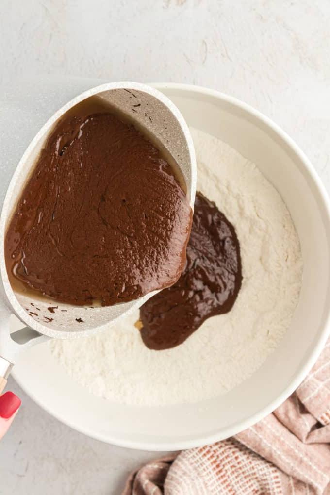 Una ciotola bianca con una miscela di farina e gli ingredienti caldi al cioccolato versati nella ciotola da una casseruola, con un panno color cuoio nell'angolo sinistro