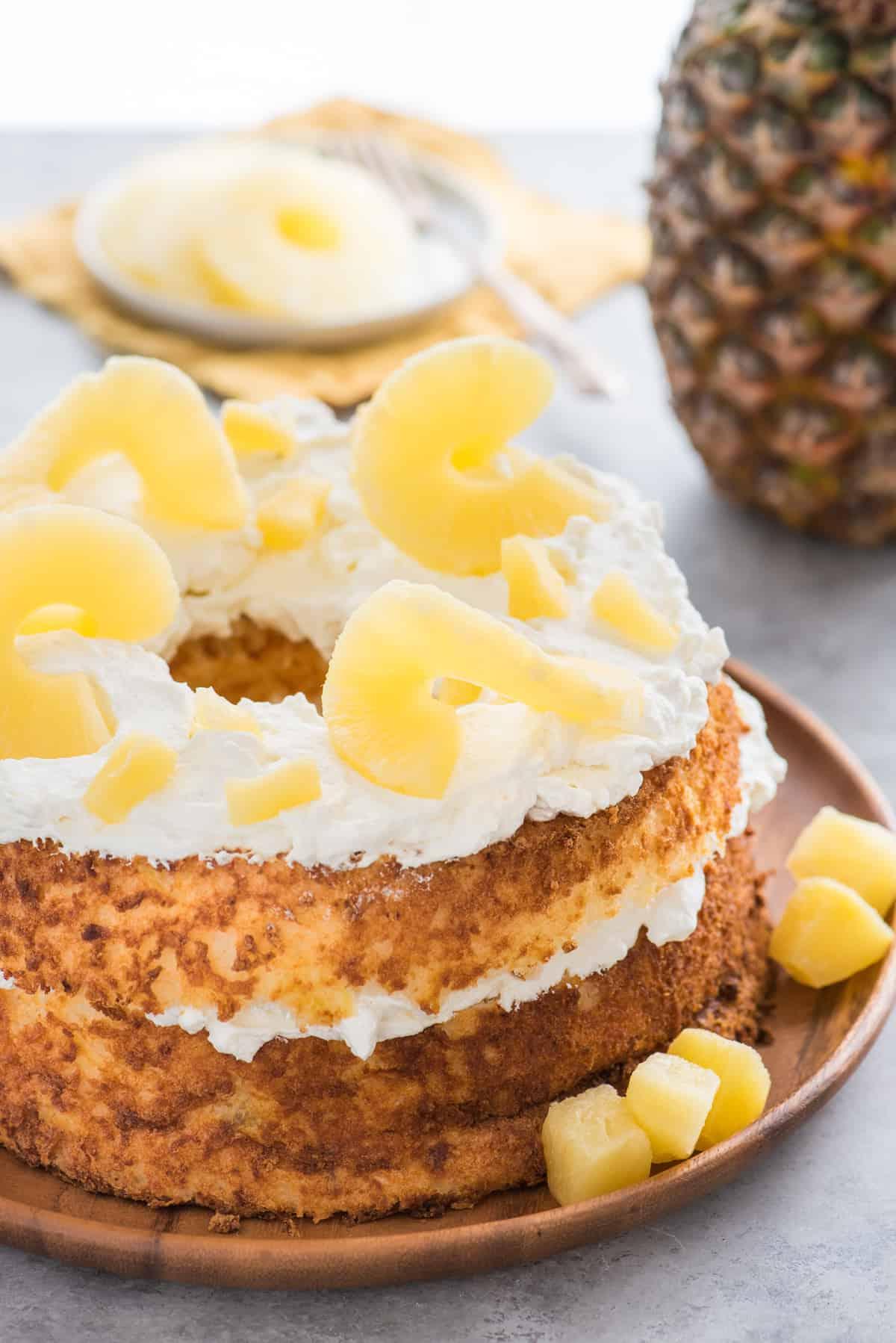 Torta Angel Food intera guarnita con panna montata e fette di ananas su vassoio di legno su sfondo bianco