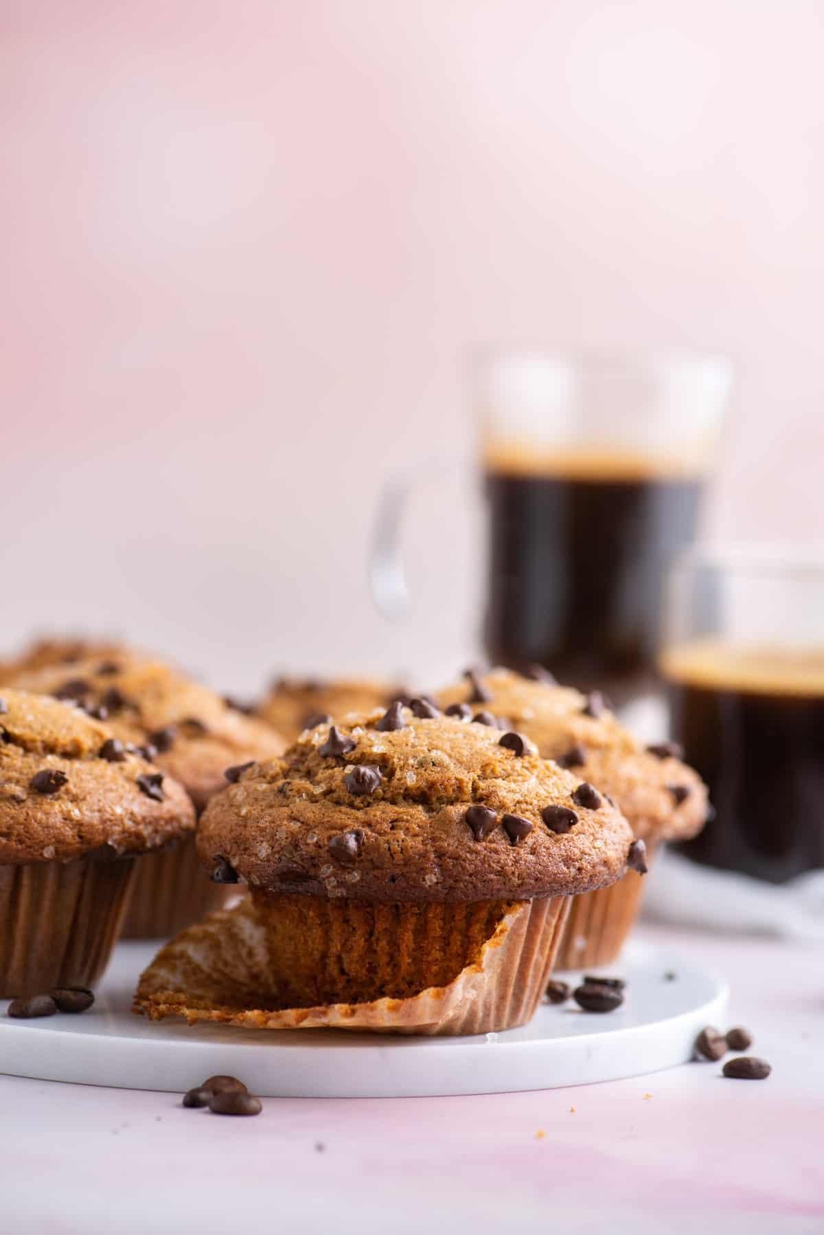 muffin al cappuccino con gocce di cioccolato su un piatto bianco, appoggiati nel loro pirottino aperto, con chicchi di caffè sparsi intorno, altri muffin e due tazze di caffè sullo sfondo