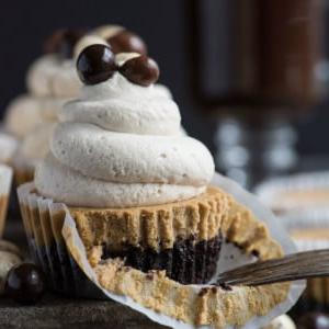 Mini Cheesecakes al Caffè 2 Una vista ravvicinata di un cupcake con base al cioccolato, crosta di cracker Graham e glassa a spirale guarnita con palline ricoperte di cioccolato. La carta parzialmente sbucciata e la forchetta richiamano l'aspetto di mini cheesecake al caffè, con altri dolcetti e una bevanda sullo sfondo.
