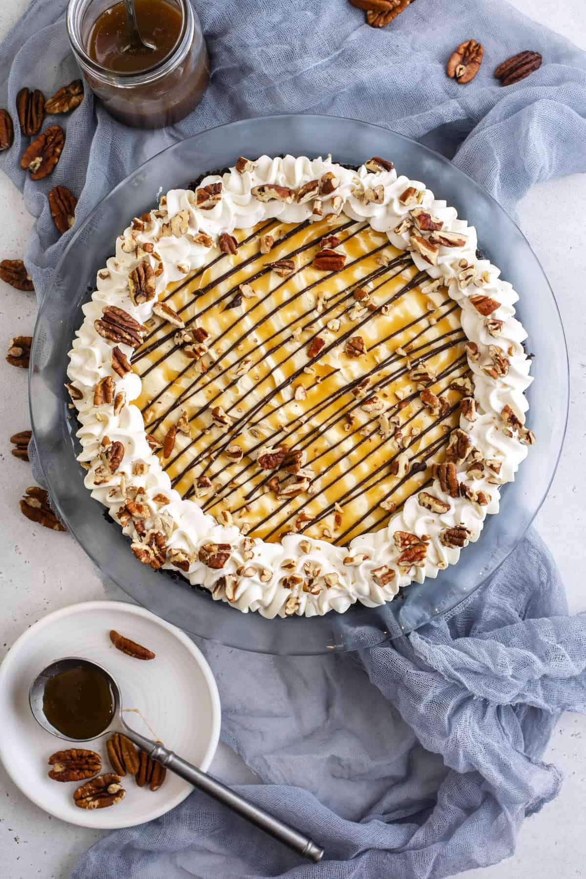 Torta di Tartaruga 8 Vista dall'alto di una Torta Tartaruga guarnita con caramello, cioccolato, panna montata e noci pecan, circondata da noci pecan e salsa al caramello.