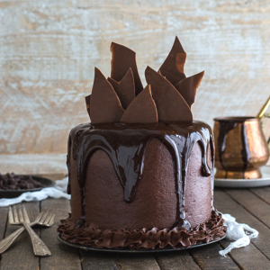 Torta al Doppio Cioccolato 4 Una torta alta al cioccolato con glassa lucida che cola dai lati, guarnita con grandi scaglie di cioccolato, su un tavolo di legno con forchette e una pentola di rame sullo sfondo.