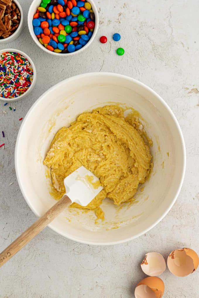 Fare Biscotti dal Mix per Torte 8 Impasto per biscotti con preparato per torte in una ciotola bianca con una spatola bianca e di legno circondata da gusci d'uovo, una ciotola di codette, una ciotola di M&M's e una ciotola di noci