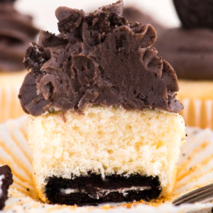 Cupcakes con base Oreo 8 Un cupcake con fondo Oreo, con impasto alla vaniglia, un biscotto al cioccolato alla base e glassa al cioccolato spessa in cima. Il cupcake è in un pirottino di carta bianco e ne è stato morso un pezzo, rivelando i suoi deliziosi strati.