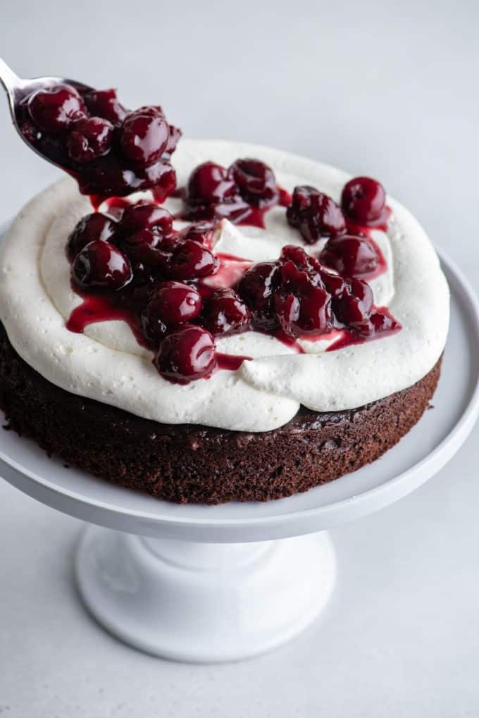 Torta Foresta Nera 8 Uno strato di pan di Spagna al cioccolato su un'alzatina bianca con la panna montata stabilizzata sopra e una salsa di ciliegie che viene cucchiaia su di essa