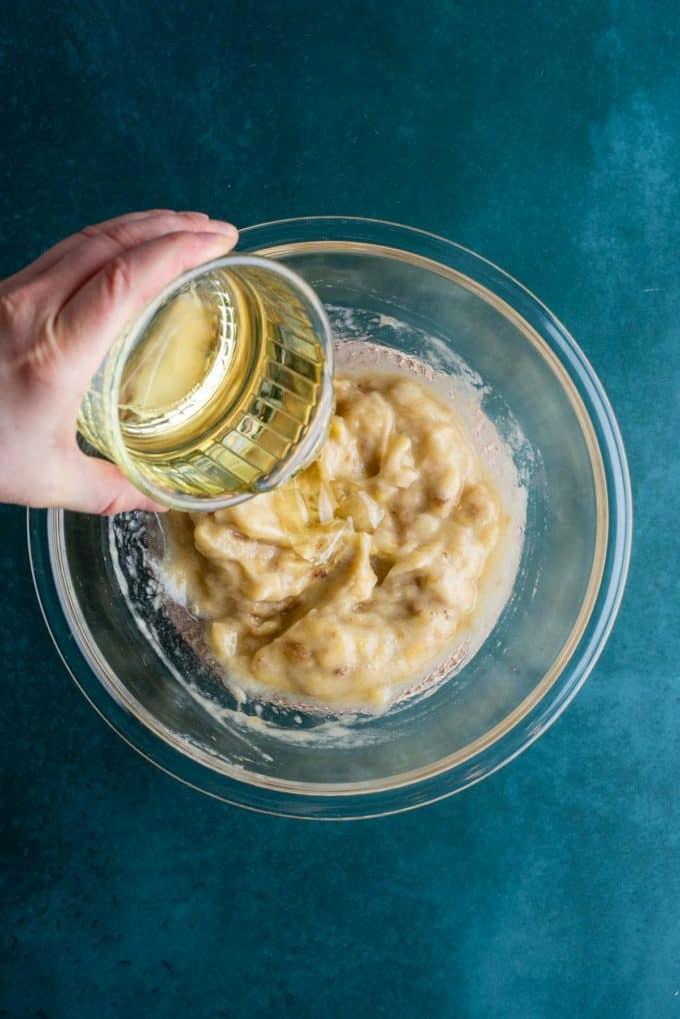 Il Miglior Pan di Banane con Gocce di Cioccolato 8 Banane schiacciate in una ciotola di vetro trasparente su una superficie color ottanio scuro con un piatto di vetro di olio vegetale che viene versato.
