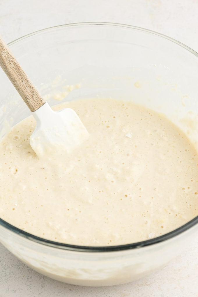 Ricetta Pancakes in Teglia 8 Impasto per pancake al forno in teglia in una ciotola di vetro trasparente su una superficie bianca con una spatola appoggiata all'interno della ciotola.