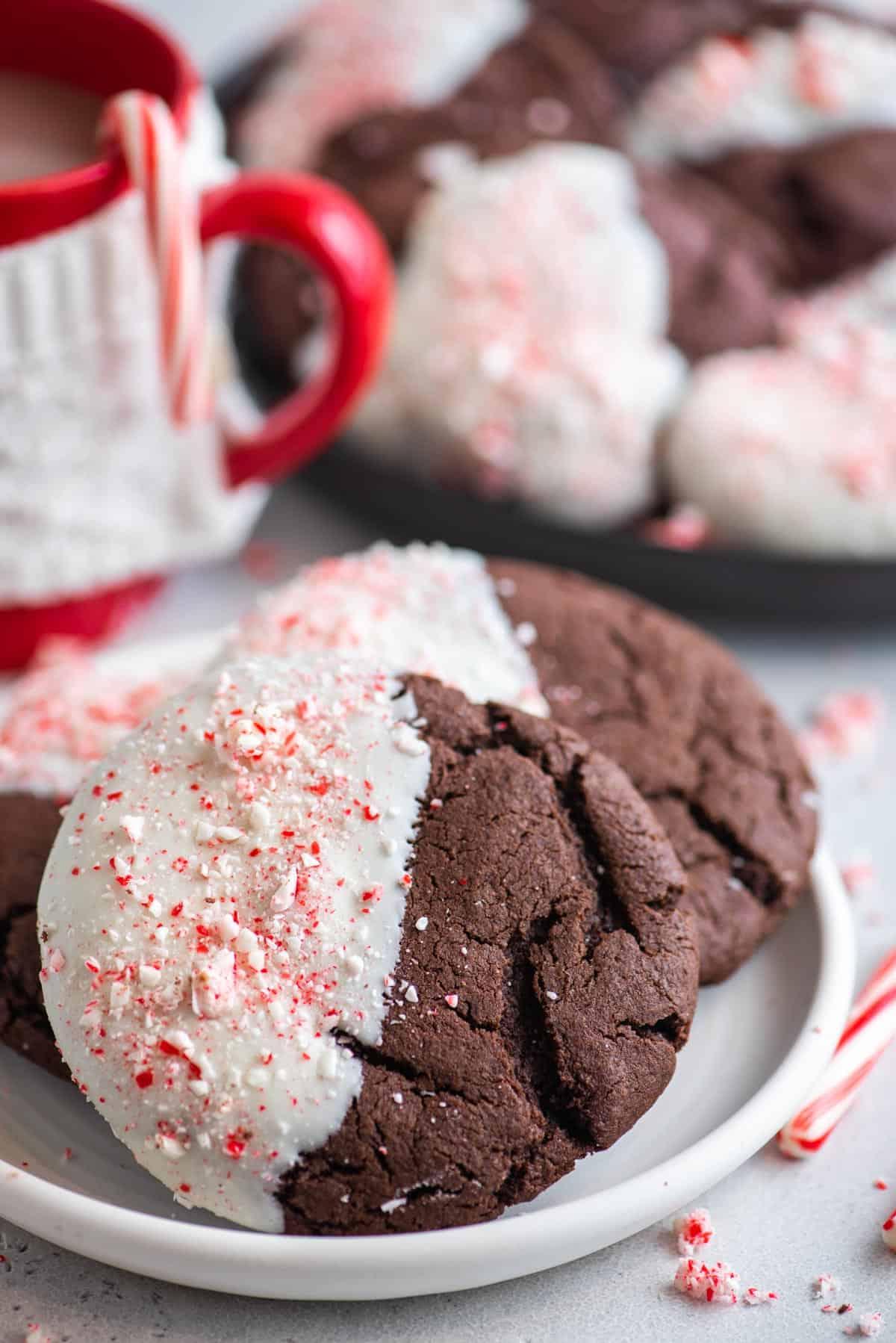 Questi Biscotti al Cioccolato e Bastoncini di Zucchero sono morbidi, gommosi, immersi nel cioccolato bianco e cosparsi di bastoncini di zucchero tritati. Biscotti al cioccolato e bastoncini di zucchero su un piatto bianco