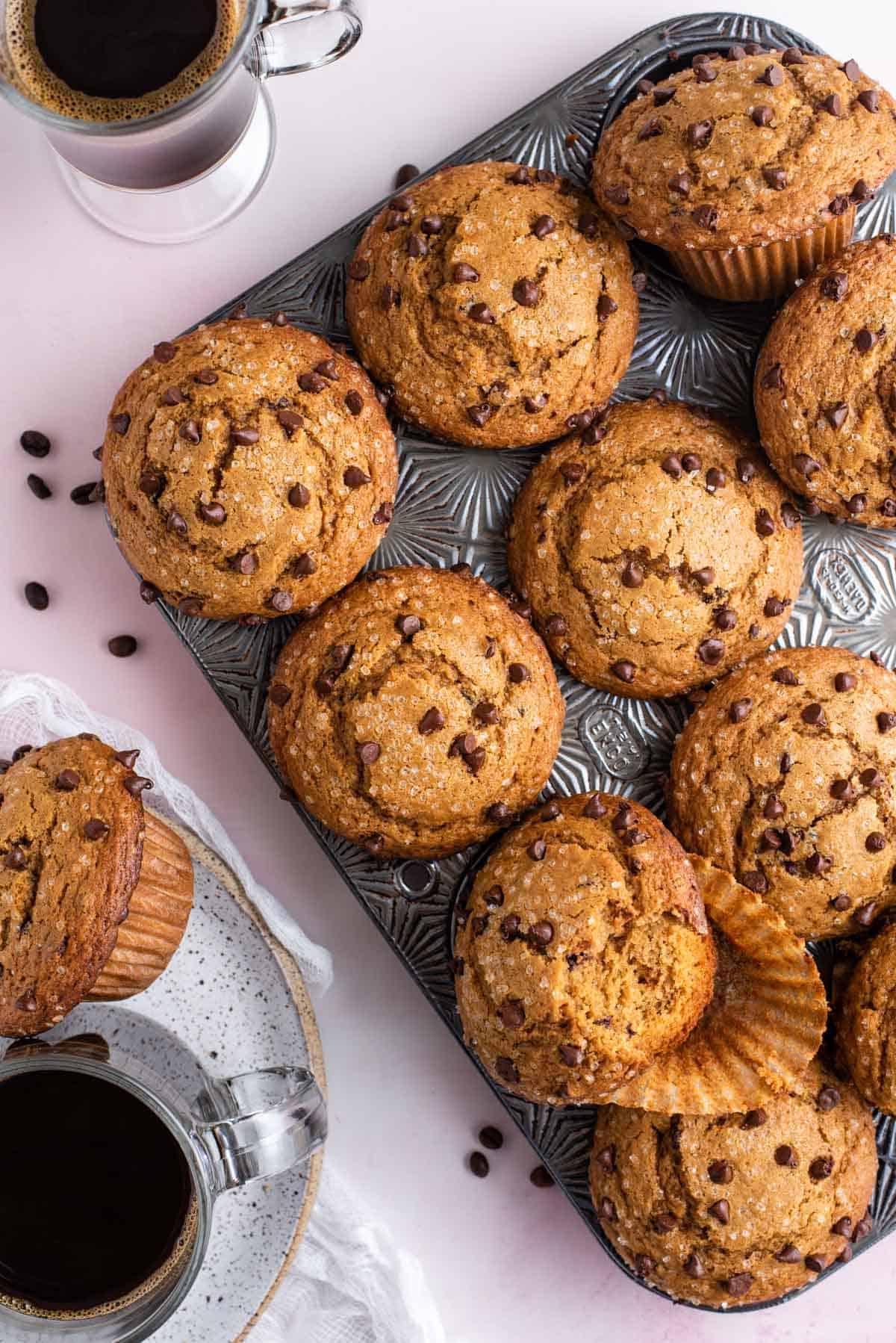 vista dall'alto di una teglia per muffin piena di muffin al cappuccino con gocce di cioccolato con chicchi di caffè sparsi intorno, con una tazza di caffè e un piatto con un muffin e una tazza di caffè accanto