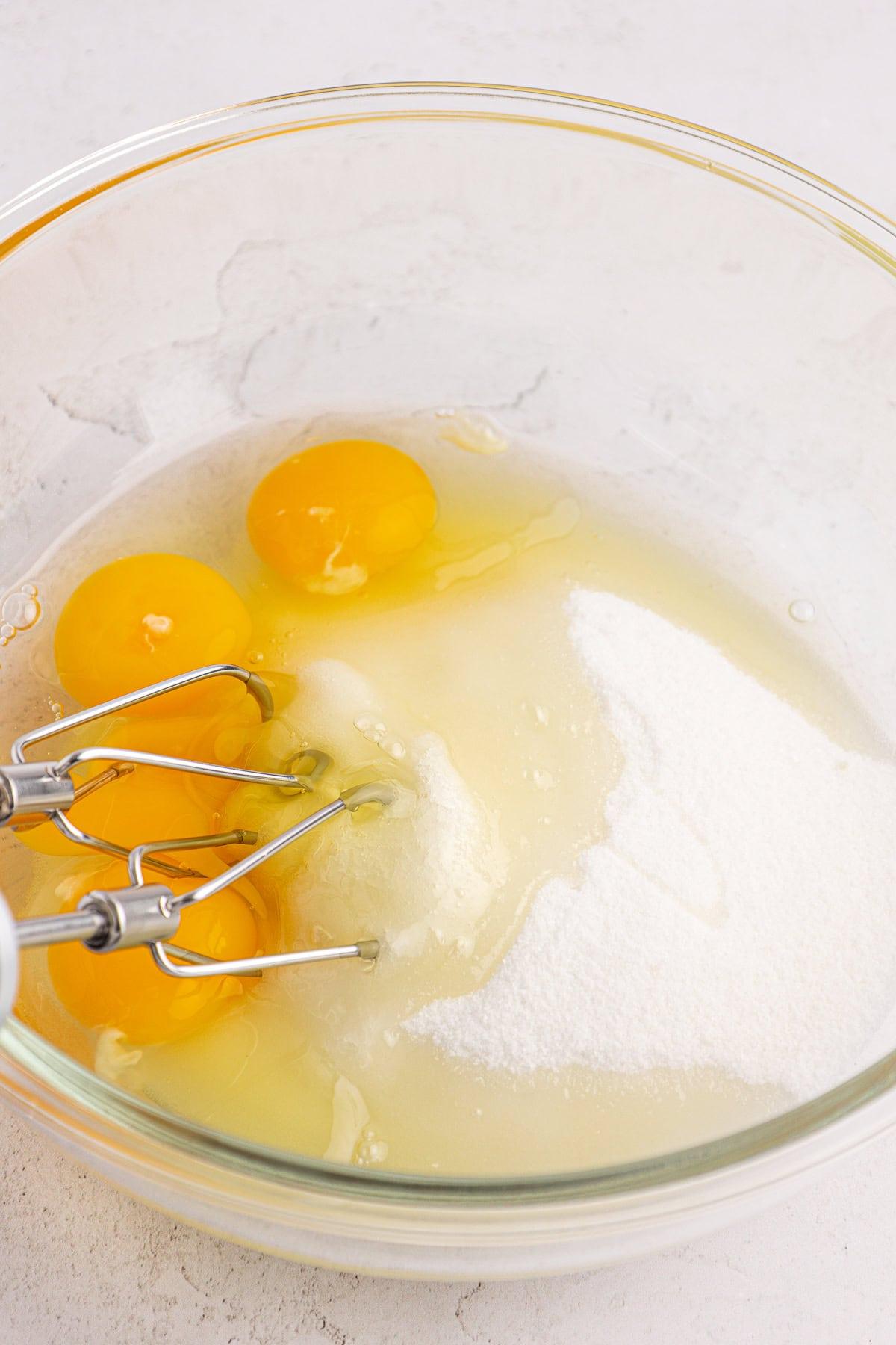 Una ciotola di vetro contenente quattro uova crude, zucchero e un frullino elettrico con le fruste metalliche, pronto per essere mischiato su un piano di lavoro chiaro.