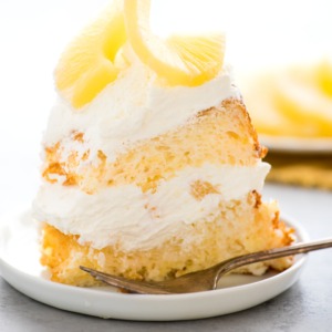 Una fetta di soffice torta Angel Food all'ananas a strati con panna montata e guarnita con fette di ananas su un piatto bianco con una forchetta accanto.