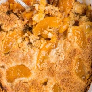 Torta Facile di Pesche con Preparato per Torta 9 Golden peach cobbler with a crumbly, browned topping and juicy peach filling in a white baking dish, partially served with a spoon—just like a homemade peach dump cake.