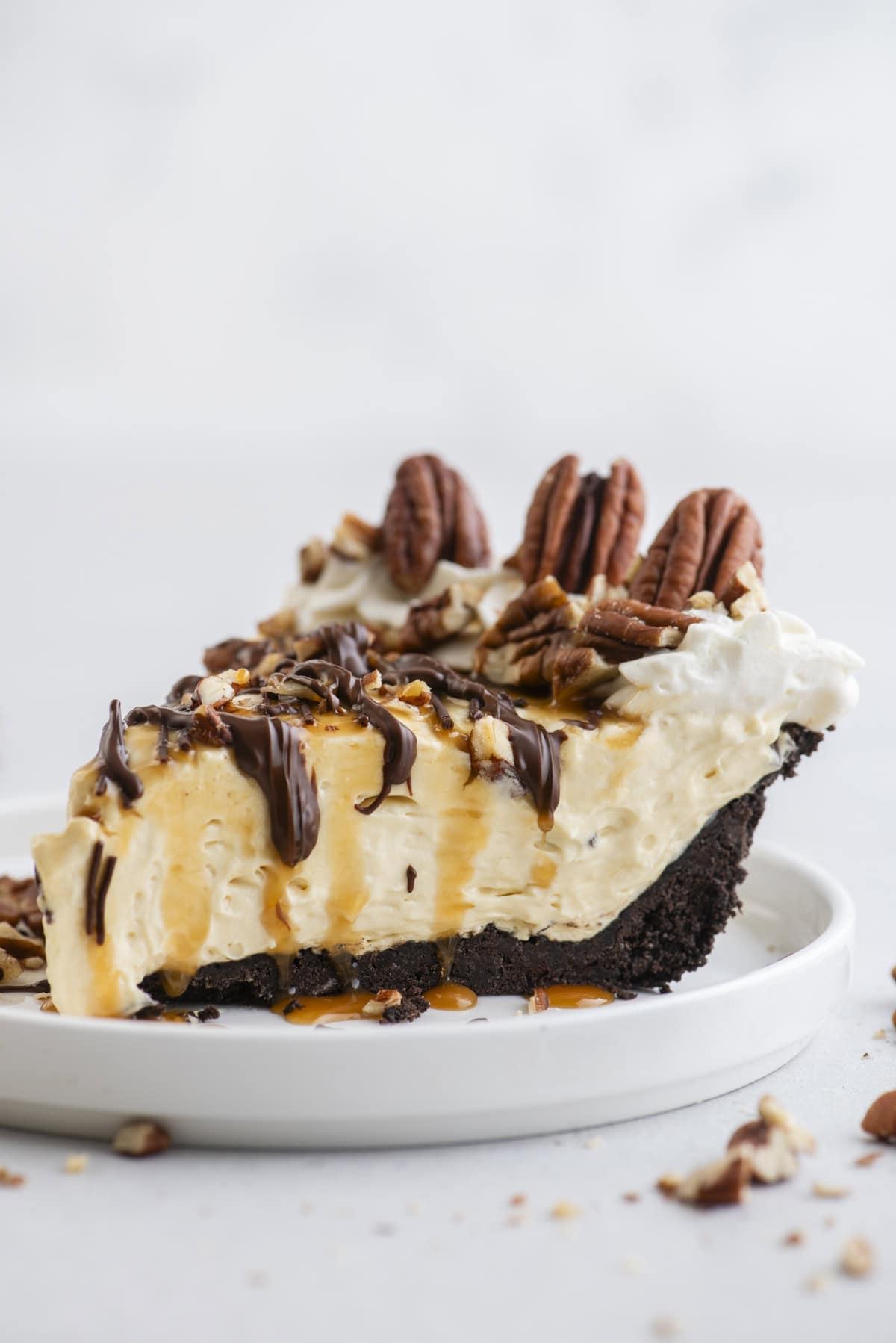Torta di Tartaruga 9 Una fetta di Torta Tartaruga guarnita con noci pecan, gocce di cioccolato e caramello, su un piccolo piatto bianco con pezzi di noci pecan sparsi intorno.