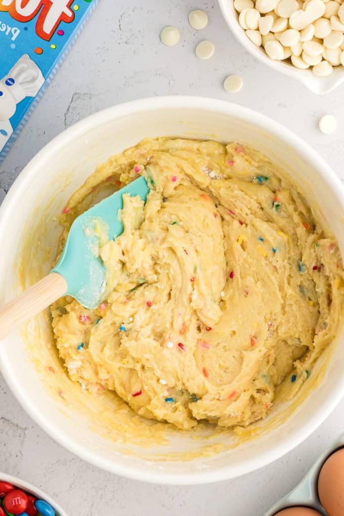 Barrette di biscotti da preparato per torte 9 a white bowl full of funfetti cake bar mix with a rubber spatula in it, a cake mix box, eggs, a bowl of M&Ms, and a bowl of white chocolate chips surrounding the bowl