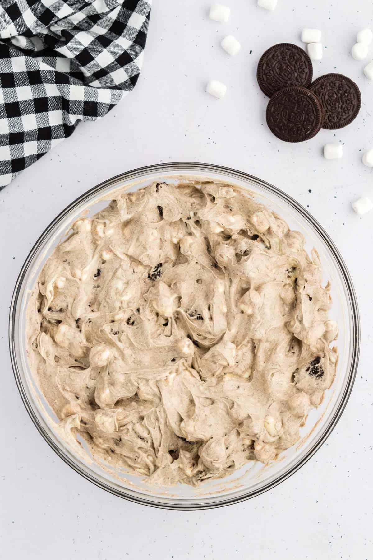 Ricetta Crema Soffice Oreo (Senza Formaggio Cremoso) 9 Fluff di Oreo in una ciotola di vetro circondato da Oreo, mini marshmallow e un panno da cucina a scacchi bianchi e neri