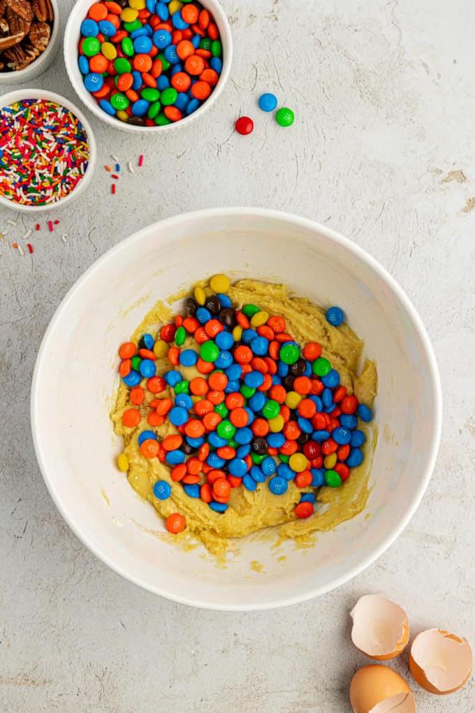Fare Biscotti dal Mix per Torte 9 Impasto per biscotti con preparato per torte in una ciotola bianca con un mucchio di M&M's sopra, circondato da gusci d'uovo, una ciotola di codette, una ciotola di M&M's e una ciotola di noci