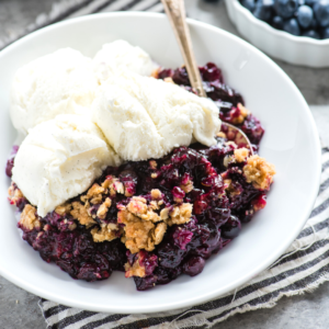 Crumble di Mirtilli Freschi 9 Una ciotola bianca di Crumble ai Mirtilli sormontata da gelato alla vaniglia, appoggiata su un panno a righe, accompagnata da un cucchiaio e una ciotola di mirtilli freschi sullo sfondo.
