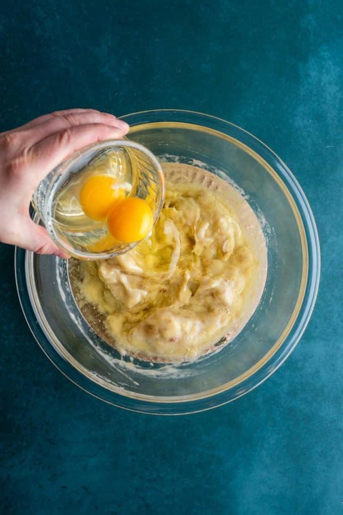 Il Miglior Pan di Banane con Gocce di Cioccolato 9 Banane schiacciate e olio vegetale in una ciotola di vetro trasparente su una superficie color ottanio scuro con due uova che vengono versate sopra.
