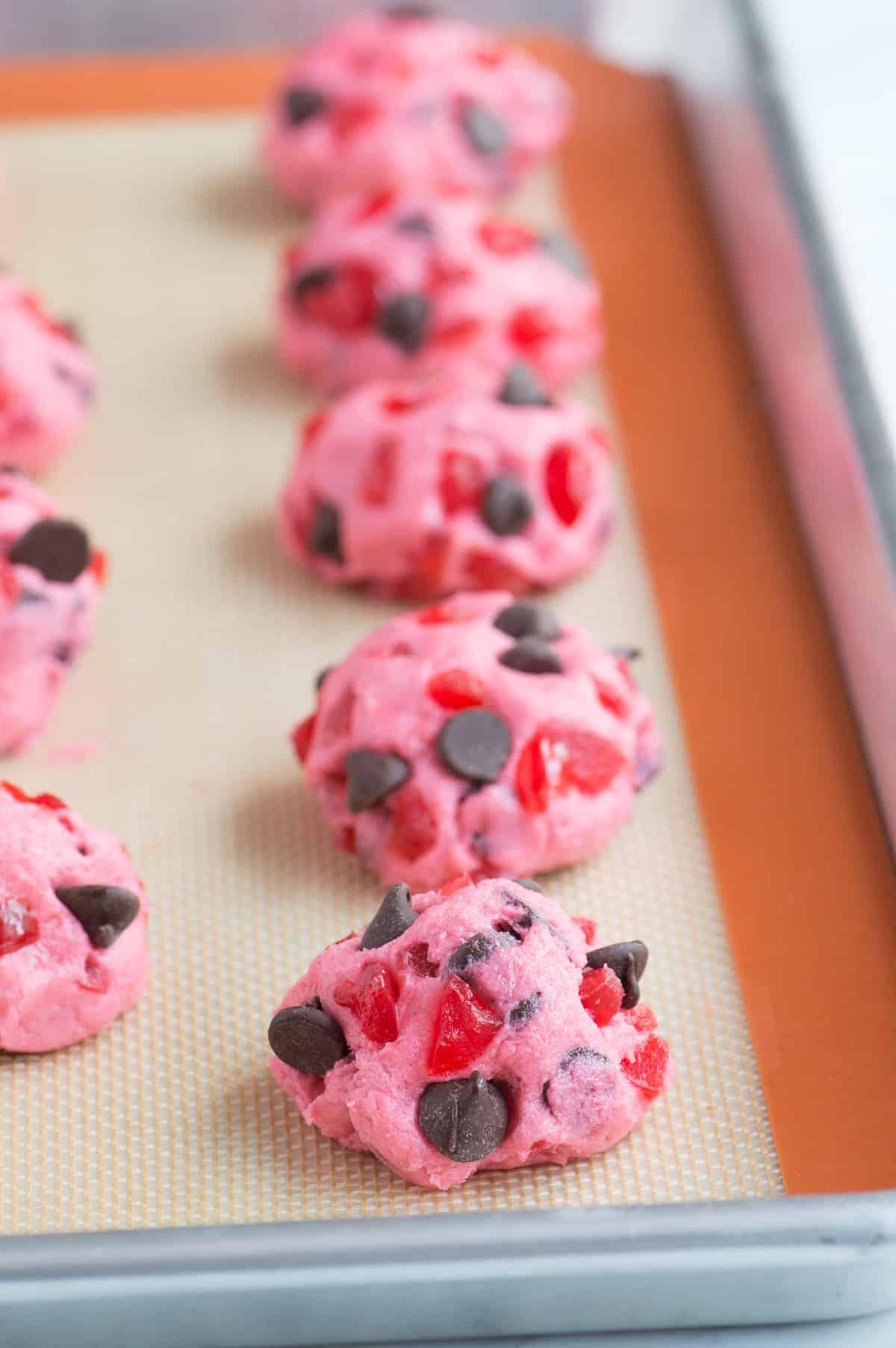 Ricetta di biscotti alle ciliegie maraschino spessi e masticabili! Ricetta di biscotti alle ciliegie candite. biscotti rosa alle ciliegie maraschino e gocce di cioccolato su teglia prima della cottura