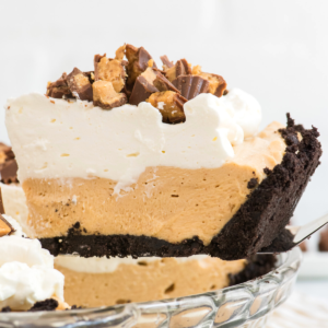 Ricetta Facile di Torta al Burro di Arachidi 9 Una fetta di cremosa torta fredda al burro di arachidi senza cottura con una crosta di biscotti al cioccolato, guarnita con panna montata e pezzi di burro di arachidi tritati, sollevata da una teglia di vetro.