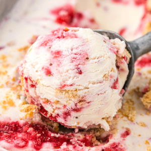 Migliore Gelato al Cheesecake alla Fragola 9 Una porzione di cremoso gelato cheesecake alle fragole con vortici di salsa rossa di lamponi e pezzetti di biscotti sbriciolati in un mestolo di metallo, circondato da altro gelato e briciole.